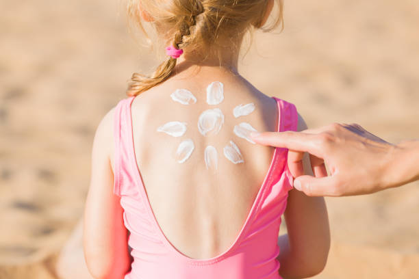Young adult mother hand applying sunscreen lotion on little girl back. Sun shape created from white cream. Skin protection. Safety sunbathing in hot sunny day at beach. Rear view. Closeup. stock photo