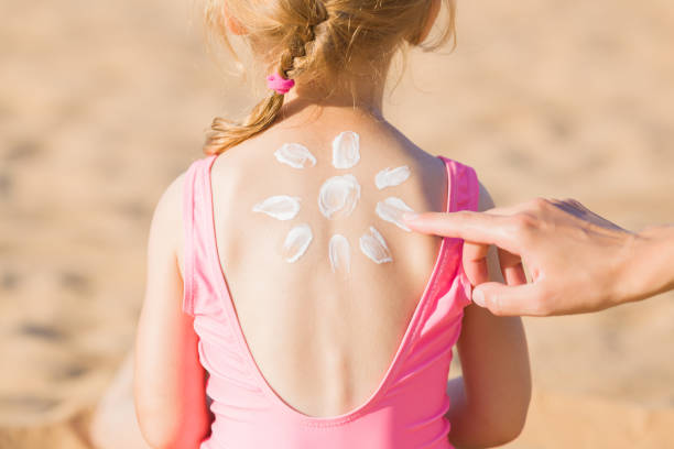 young adult mother hand applying sunscreen lotion on little girl back. sun shape created from white cream. skin protection. safety sunbathing in hot sunny day at beach. rear view. closeup. - sky human hand water white imagens e fotografias de stock