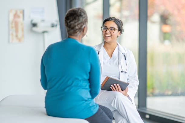 Doctor Meeting with a Senior Patient A senior patient of African decent, sits up on an exam table across from her female doctor as they discuss her medical concerns.  She is dressed casually and listening attentively as the doctor talks with her.  Her female doctor is wearing a white lab coat and has a tablet in her hands as she takes notes throughout. general practitioner stock pictures, royalty-free photos & images