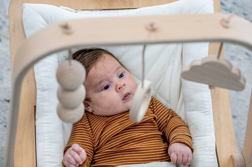A sweet little newborn baby boy, lays in a swing as he focuses on the mobile above him.  He is dressed casually in a striped shirt and is looking wide-eyed on wonder.