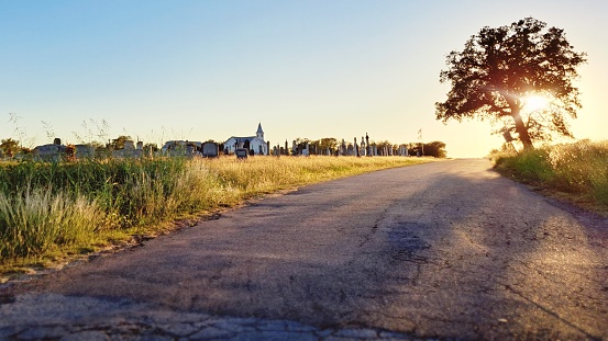 Approaching sunset along a rural central Texas road, where a quaint white church and its cemetery can be seen on the horizon.