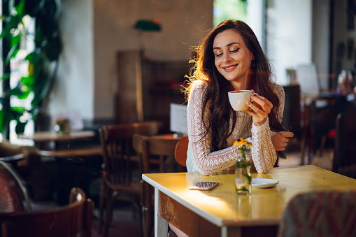 Portrait of beautiful young woman drinking a cup of tea in cafeteria. Happy female in a cafe drinking coffee with her eyes closed while daydreaming. Smiling lady relaxing in a coffee shop. Copy space.