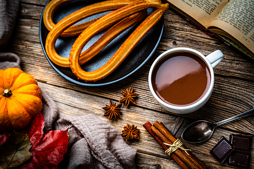 Sweet food: churros and hot chocolate mug shot from above on rustic wooden table. Dark chocolate bar and pieces, cinnamon sticks, star anise, an open book and autumn leaves complete the composition. High resolution 42Mp studio digital capture taken with SONY A7rII and Zeiss Batis 40mm F2.0 CF lens