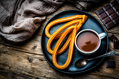 Sweet food: churros and hot chocolate mug shot from above on rustic wooden table. Dark chocolate bar and pieces complete the composition. High resolution 42Mp studio digital capture taken with SONY A7rII and Zeiss Batis 40mm F2.0 CF lens