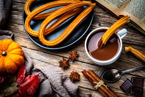 Sweet food: churros and hot chocolate mug shot from above on rustic wooden table. Dark chocolate bar and pieces, cinnamon sticks, star anise, an open book and autumn leaves complete the composition. High resolution 42Mp studio digital capture taken with SONY A7rII and Zeiss Batis 40mm F2.0 CF lens