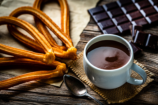 Sweet food: churros and hot chocolate mug shot on rustic wooden table. Dark chocolate bar and pieces complete the composition. High resolution 42Mp studio digital capture taken with Sony A7rII and Sony FE 90mm f2.8 macro G OSS lens
