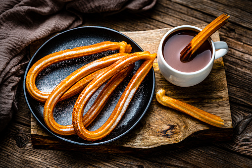 Sweet food: churros and hot chocolate mug shot from above on rustic wooden table. Dark chocolate bar and pieces complete the composition. High resolution 42Mp studio digital capture taken with Sony A7rII and Sony FE 90mm f2.8 macro G OSS lens