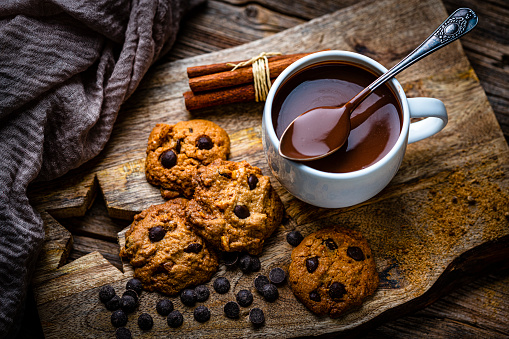 Sweet food: homemade chocolate chip cookies and hot chocolate mug shot from above on rustic wooden table. Dark chocolate bar and pieces, cinnamon sticks, star anise, an open book and autumn leaves complete the composition. High resolution 42Mp studio digital capture taken with SONY A7rII and Zeiss Batis 40mm F2.0 CF lens