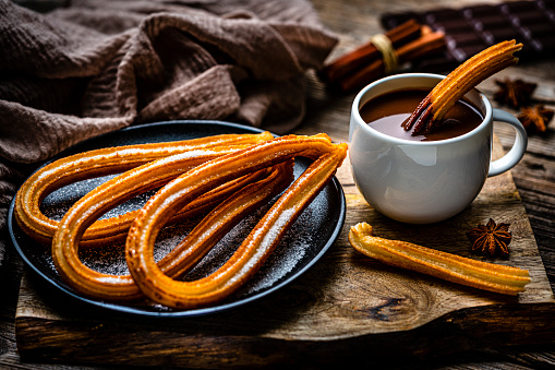Sweet food: churros plate and hot chocolate mug shot on rustic wooden table. Dark chocolate bar and pieces complete the composition. High resolution 42Mp studio digital capture taken with Sony A7rII and Sony FE 90mm f2.8 macro G OSS lens