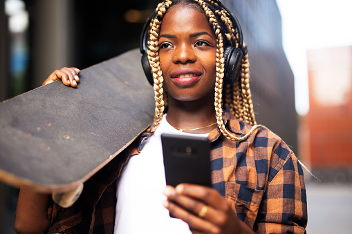 African woman with skateboard. Beautiful urban girl using the phone outdoors.