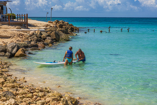 Aruba. Oranjestad. 09.12.2022. Beautiful view of couple fixing surfboard in water on coastline.