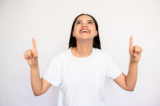 Portrait of happy young woman pointing fingers upwards over white background. Caucasian lady wearing white T-shirt looking up and smiling. Advertising concept