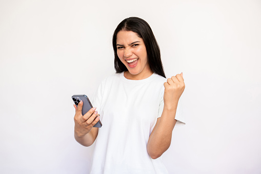 Portrait of happy young woman with phone making winning gesture over white background. Excited Caucasian lady wearing white T-shirt celebrating success. Good news concept
