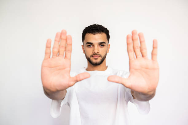 Portrait of confident young man making restriction sign over white background. Bearded man wearing white T-shirt stretching arms in stop gesture. Forbiddance concept