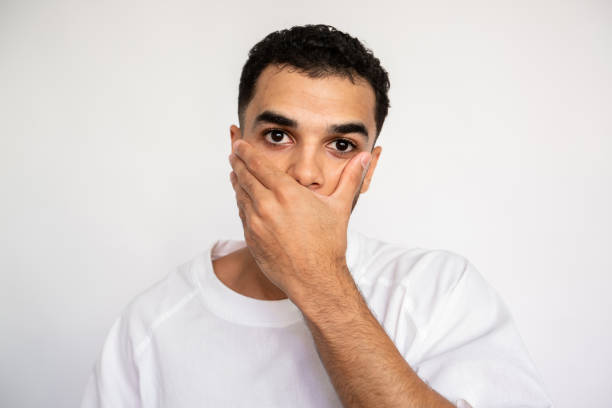 Portrait of worried young man covering mouth with hand Portrait of worried young man covering mouth with hand over white background. Bearded man wearing white T-shirt looking at camera in fear or stress. Fault or problem concept speak no evil stock pictures, royalty-free photos & images