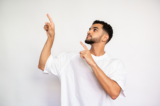 Portrait of serious young man pointing forefingers upwards over white background. Bearded man wearing white T-shirt looking up and showing something. Advertising concept