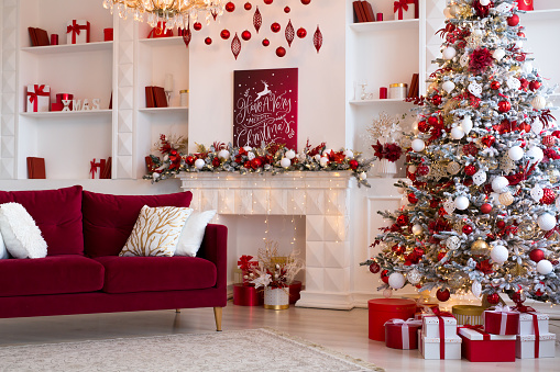 Interior of bright modern living room with fireplace, chandelier and comfortable sofa decorated with Christmas tree and red gifts.