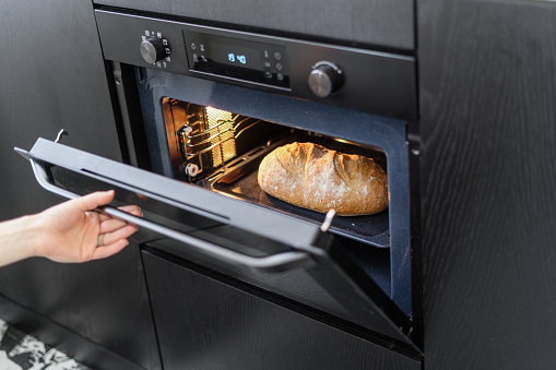cooking at home, cropped shot of female hand opening cooker door with homemade loaf bread inside on tray, modern technology appliance