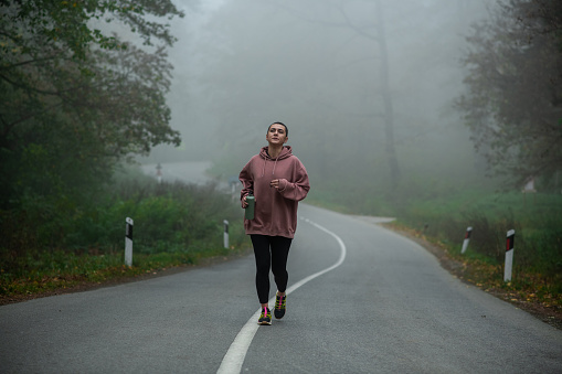 Young female athlete on a morning run on a foggy forest road. Beautiful young woman on her jog, wearing sportswear and drinking water from her eco bottle.