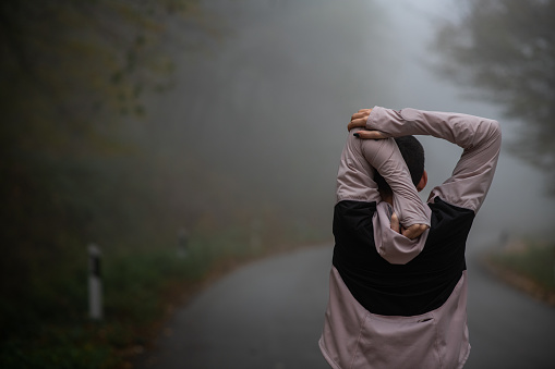 Young female athlete on a morning run on a foggy forest road. Beautiful, determined young woman on her jog, wearing sportswear. Focused young woman with short hair jogging and stretching.