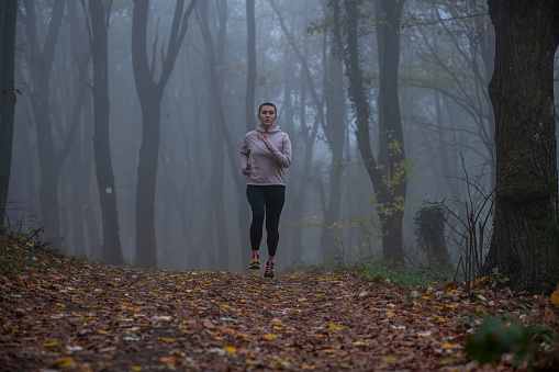 Young female athlete on a morning run on a foggy forest road. Beautiful, determined young woman on her jog, wearing sportswear. Focused young woman with short hair jogging.
