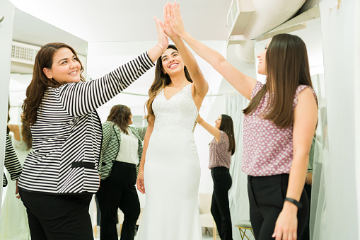 Cheerful friends and future bride woman making a high five and celebrating while shopping for a wedding dress