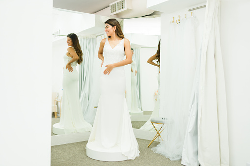 Attractive latin woman buying a beautiful wedding dress and looking in the mirror at the dressing room