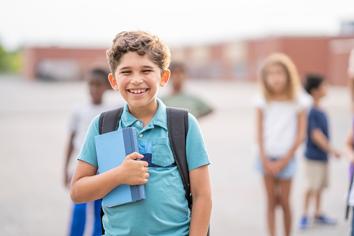A young Elementary schoolboy stands outside as he poses for a portrait in front of his school.  He is dressed casually in shorts and a t-shirt and is holding his books tightly as his peers stand in the background.