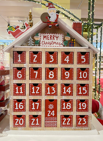 an advent calendar in the shape of a little house with the numbers of the days of December in small drawers, christmas calendar decorated with santa claus and elves and a merry christmas message, vertical