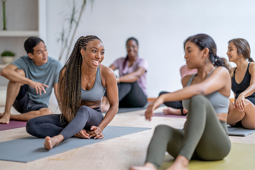 A small group of mature adults sit on individual yoga mats as they take a break after their fitness class.  They are each dressed casually in athletic wear and are smiling as they talk amongst themselves.
