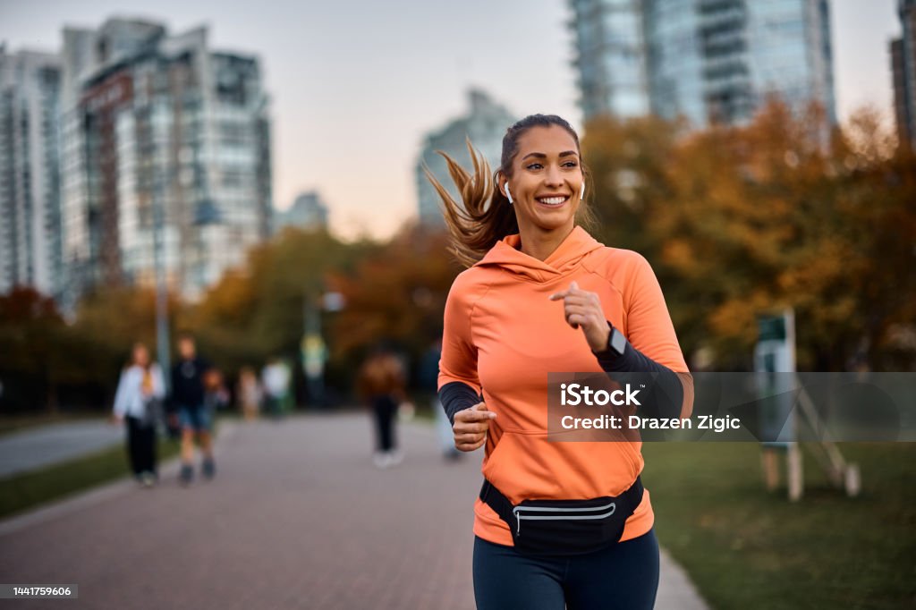 Happy sportswoman with earbuds running in the park. Young happy athletic woman listening music on earphones while jogging in the park. Running Stock Photo