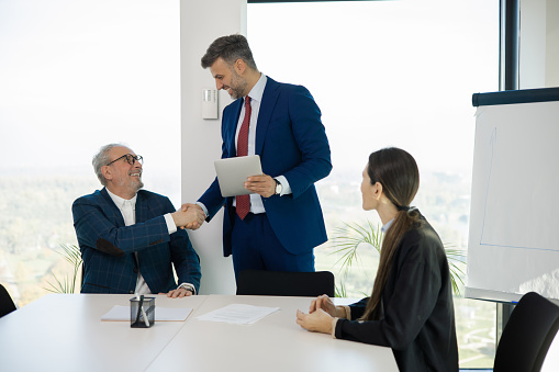 senior confident male speaker in elegant gray suit explaining his opinion to partners, other executives at multi-ethnic meeting in office, using microphones for giving speech