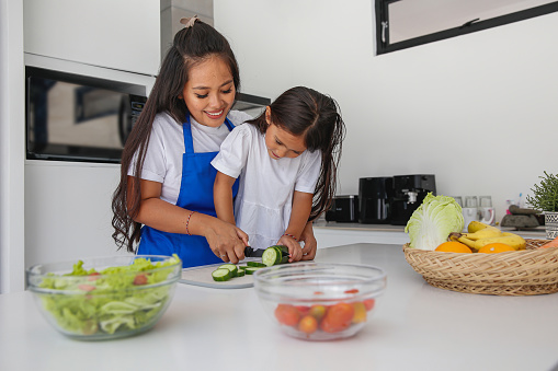 Close-up shot of mother teaching little daughter how to cut cucumber with a kitchen knife.