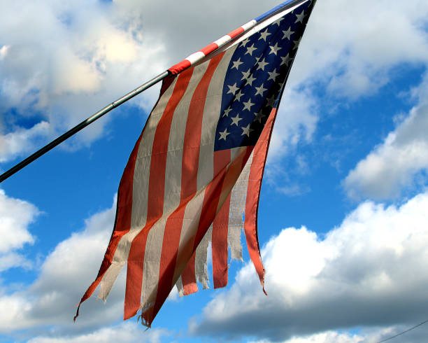 Tattered US flag in front of a blue sky with clouds.