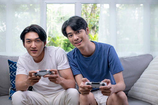 Asian gay lgbt men couple play video games at home, males having funny happy moment together on sofa in living room.