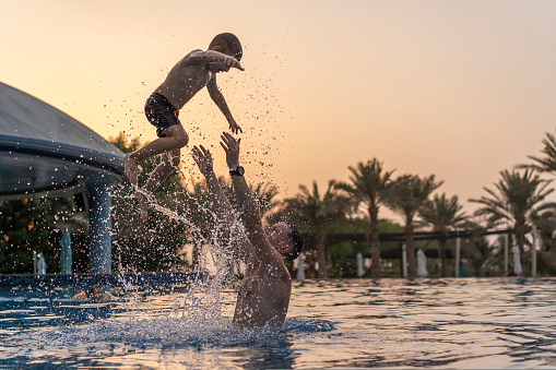 Dad and son are playing in the swimming pool. The father tosses the son up. Sunset in the United Arab Emirates. International father's day