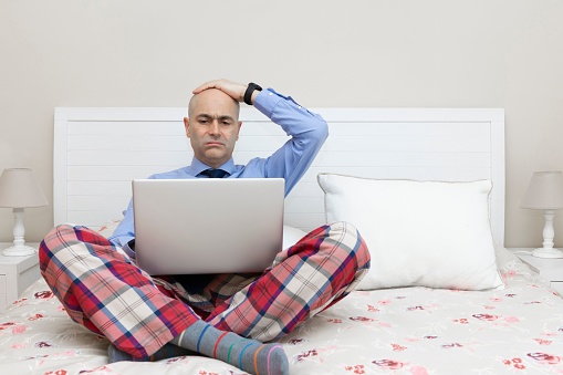 Man working with a laptop on a bed dressed in a shirt and tie and pajama pants and with his hand over his head with a worried expression.