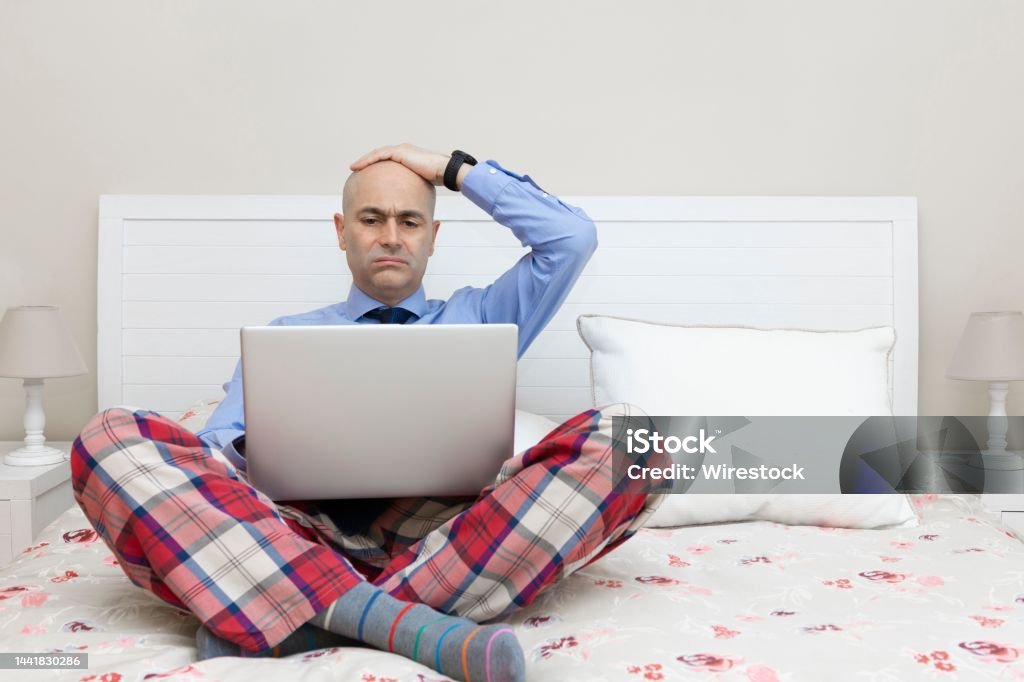 Man working with a laptop on a bed dressed in a shirt and tie with a worried expression Man working with a laptop on a bed dressed in a shirt and tie and pajama pants and with his hand over his head with a worried expression. Pajamas Stock Photo