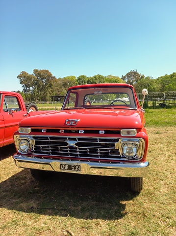 San Isidro, Argentina – October 08, 2022: Old red 1968 Ford F 100 pickup truck in the countryside. Nature, grass, trees. Autoclasica 2022 classic car show. Front view. Grille