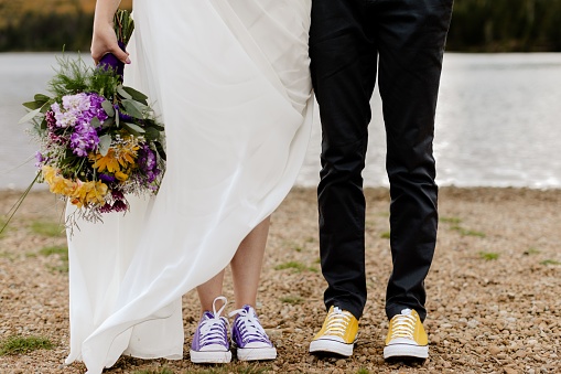 A groom in yellow sneakers standing by the bride in purple sneakers holding a purple-yellow bouquet