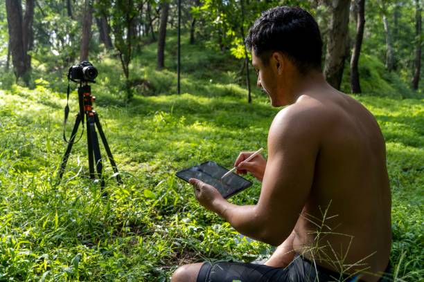 istock Young male drawing on a tablet sitting on fresh grass in a forest 1441839527