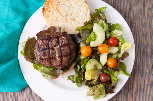 A top view shot of a delicious garlic pork burger with a green salad on the size in a white plate on a wood surface