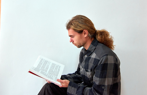 A handsome Caucasian man with long hair reading a book on the background of the white wall