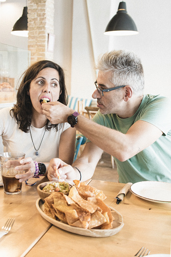 Adult couple enjoying plate of nachos with spicy green sauce together, sitting at restaurant table. Tourist couple having good time in small eatery, enjoying food and each other company.