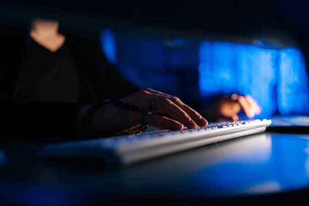 Close-up hands of unrecognizable hacker man working typing on keyboard laptop computer sitting at desk in dark room with blue neon lights.