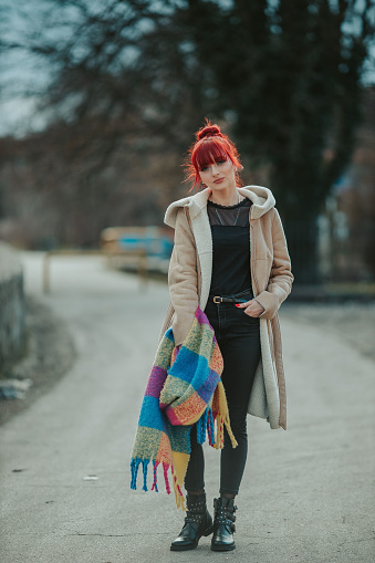 A full body shot of a red-haired girl with bangs strolling in the city with a warm winter coat holding a colorful scarf