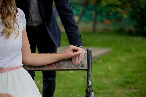 A closeup of a newly married couple holding hands on a bench in a park with a blurry background