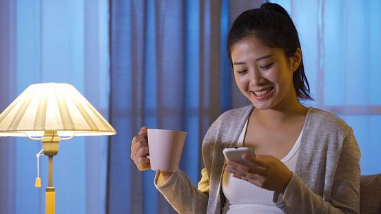 portrait cheerful young woman phubbing at home in the evening. asian girl holding smarthpone chuckling over interesting posts on social media. modern technology and real moments.