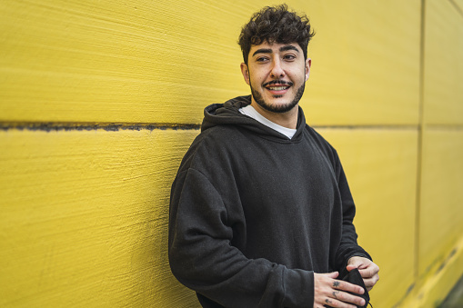 A handsome smiling European skater in hoodie posing in front of a yellow wall with facial mask in hands - new normal concept