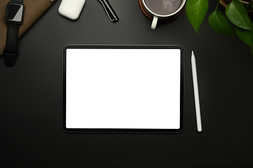Digital tablet with white empty display, stylus pen, coffee cup and notepad on black wooden table.